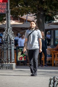 Фото тату: Мужчина с длинными волосами и татуировкой ворона на предплечье, идущий по улице в солнечный день.