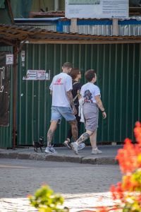 Фото мужчины с татуировкой на руке, идущего по улице в футболке. Вид сбоку, солнечный день.