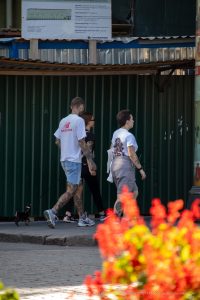 Фото мужчины с татуировкой на руке, идущего по улице в футболке. Вид сбоку, солнечный день.