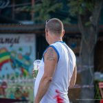 Фото татуировки на плече мужчины, стоящего на фоне яркой, красочной стены. Рисунок татуировки выполнен в темных тонах, с деталями и контурами.
