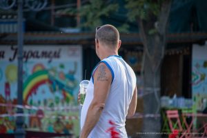 Фото татуировки на плече мужчины, стоящего на фоне яркой, красочной стены. Рисунок татуировки выполнен в темных тонах, с деталями и контурами.