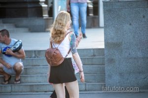 Фотография девушки с акварельной татуировкой на руке. Татуировка изображает цветы и маску, выполнена в ярких цветах. Девушка спускается по лестнице.