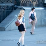 Фотография девушки с акварельной татуировкой на руке. Татуировка изображает цветы и маску, выполнена в ярких цветах. Девушка идет по улице.