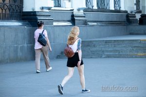 Фотография девушки с акварельной татуировкой на руке. Татуировка изображает цветы и маску, выполнена в ярких цветах. Девушка идет по улице.