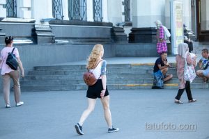 Фотография девушки с акварельной татуировкой на руке. Татуировка изображает цветы и маску, выполнена в ярких цветах. Девушка идет по улице.