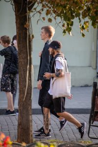 Фото мужчины с татуировкой на руке, идущего по улице с сумкой. Вид сбоку, солнечный день.