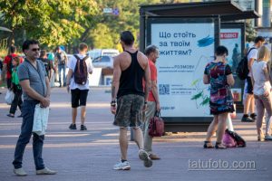 Фотография мужчины с татуировкой на руке, стоящего рядом с автобусной остановкой. Татуировка выполнена в темных тонах и занимает большую часть предплечья.