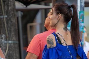 Фото тату на шее и плече девушки в синем платье. Видны надписи и рисунок черепа, выполненные в реалистичном стиле. Уличная татуировка.