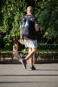 Фото мужчины с татуировками на шее и ногах, идущего по парку с рюкзаком. Вид сзади.