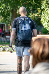 Фото тату на теле мужчины: татуировки на шее, предплечье и ногах, выполненные в разных стилях. Мужчина с рюкзаком на улице.