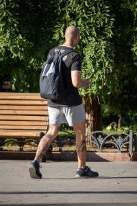 Фото мужчины с татуировками на шее и ногах, идущего по парку с рюкзаком. Вид сзади.