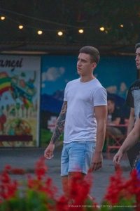 Фотография мужчины с татуировкой на руке, стоящего на фоне яркой афиши. Татуировка выполнена в темных тонах и занимает большую часть предплечья.