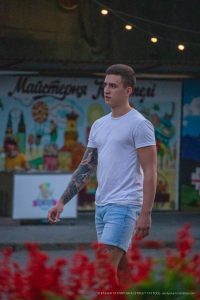 Фотография мужчины с татуировкой на руке, стоящего на фоне яркой афиши. Татуировка выполнена в темных тонах и занимает большую часть предплечья.