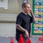 Мужчина с бритой головой и татуировками на руках и ногах идет по улице, разговаривая по телефону.