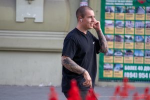Мужчина с бритой головой и татуировками на руках и ногах идет по улице, разговаривая по телефону.