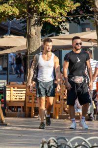 Фотография двух мужчин, один с татуировкой на предплечье, идущих по улице в солнечный день. Один одет в белую майку, другой в черную футболку.