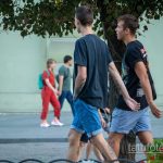 Фотография двух мужчин, идущих по улице, с видимыми татуировками на руках. Летний день, городская сцена.