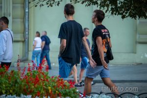Фотография двух мужчин, идущих по улице, с видимыми татуировками на руках. Летний день, городская сцена.