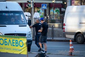 Фото тату: мужчина с татуировкой на плече стоит возле белого фургона, на фоне городской улицы.