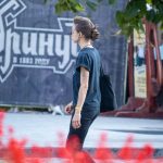 Фотография девушки с татуировкой на предплечье, идущей по улице. На заднем плане видна вывеска и другие прохожие.