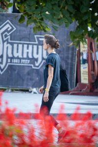 Фотография девушки с татуировкой на предплечье, идущей по улице. На заднем плане видна вывеска и другие прохожие.