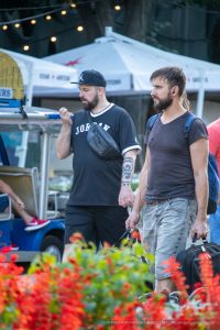 Фотография мужчины с татуировкой на предплечье, идущего с другим мужчиной на фоне городской улицы с яркими цветами.