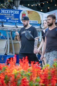 Фотография мужчины с татуировкой на предплечье, идущего с двумя девушками на фоне городской улицы с яркими цветами и архитектурой.