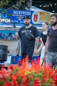 Фотография мужчины с татуировкой на предплечье, идущего с двумя девушками на фоне городской улицы с яркими цветами.