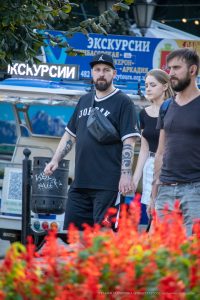 Фотография мужчины с татуировкой на предплечье, стоящего на фоне городской улицы с яркими цветами и архитектурой.