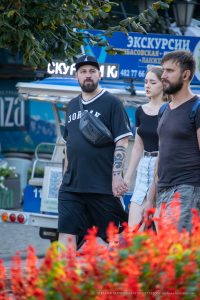 Фотография мужчины с татуировкой на предплечье, идущего с двумя девушками на фоне городской улицы.
