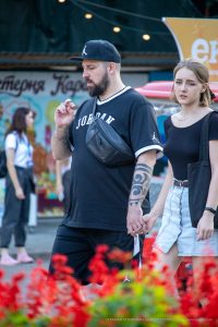 Фотография мужчины с татуировкой на предплечье, идущего рядом с девушкой на фоне городской улицы с яркими цветами.