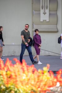 Фото мужчины с татуировкой ворона на предплечье, идущего по улице с женщиной на фоне красных цветов. Детализированный рисунок татуировки.