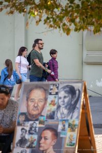 Фото мужчины с татуировкой ворона на предплечье, стоящего рядом с художниками, рисующими портреты на улице. Детализированный рисунок татуировки.