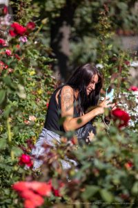 Фотография девушки с татуировкой на плече, стоящей среди цветущих роз. Татуировка выполнена в темных тонах и изображает животное.