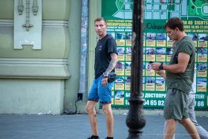 Фотография мужчины с татуировкой на руке, идущего по улице. Татуировка выполнена в черном цвете и покрывает большую часть предплечья. Уличная фотография.