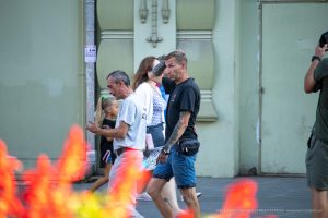 Фото тату на руке мужчины, идущего по улице. Татуировка выполнена в черном цвете и покрывает большую часть предплечья. Уличная фотография.