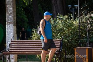 Фото мужчины с татуировкой волка на плече, идущего по улице с рюкзаком. Черно-серый рисунок татуировки.