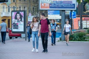 Фотография пары, идущей по улице города. Мужчина в красной и черной футболке с татуировкой на руке, девушка в белой футболке и джинсах.