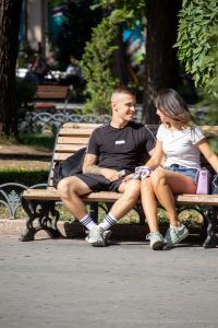 Фотография пары, сидящей на скамейке в парке. У парня заметна татуировка на предплечье, выполненная в темных тонах.