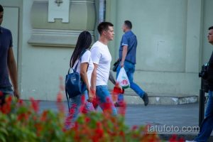 Фотография пары, идущей по улице города. Мужчина в белой футболке и джинсах, девушка в рюкзаке и джинсах. На заднем плане другие прохожие и городская архитектура.