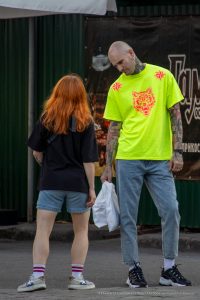 Фото татуировки на руке мужчины, изображающей ворона с красными акцентами. Тату выполнено в ярких цветах, с высокой детализацией. Виден городской фон.
