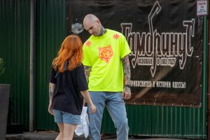 Фото татуировки на руке мужчины, изображающей ворона с красными акцентами. Тату выполнено в ярких цветах, с высокой детализацией. Виден городской фон.