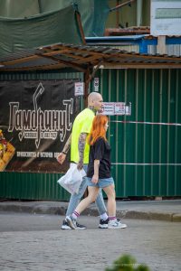 Фото татуировки на руке мужчины, изображающей ворона и сложные узоры. Тату выполнено в черном и красном цветах, с высокой детализацией. Виден городской фон.