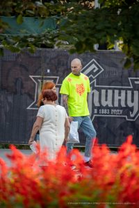Фото тату: Мужчина в яркой лаймовой футболке с татуировкой волка на плече и рукаве, идущий рядом с женщиной на фоне красных цветов.