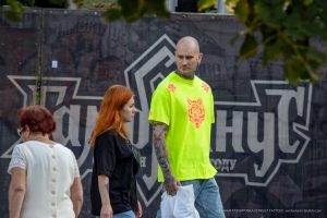 Фото тату: Мужчина в яркой лаймовой футболке с татуировкой волка на плече и рукаве, идущий рядом с девушкой.