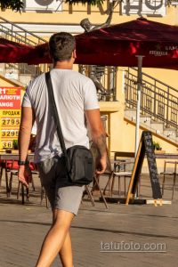 Фото мужчины с татуировкой на руке, идущего по улице. Видна часть татуировки с темными контурами. На плече сумка через плечо.