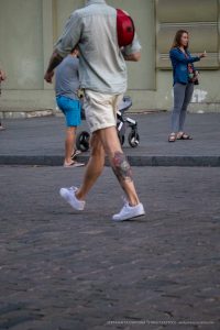 Фотография татуировки на икре мужчины, идущего по улице. Тату выполнено в ярких цветах, с детальной проработкой элементов. Виден городской фон и прохожие.