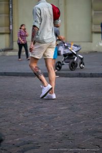 Фото тату на икре мужчины, идущего по улице. Рисунок выполнен в ярких цветах, с преобладанием красного и черного. Видны детализированные элементы рисунка.
