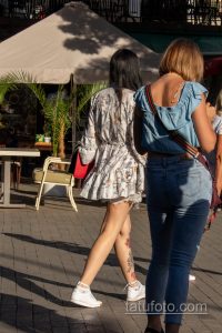 Фотография двух женщин, прогуливающихся по улице, с акцентом на ногу одной из них, украшенную татуировкой с цветами и красными мазками.