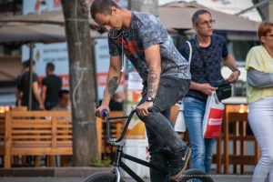 Фото БМХ-райдера с татуировками на руках, выполняющего трюк на велосипеде. Видны детализированные рисунки татуировок, включая ворона и черепа.